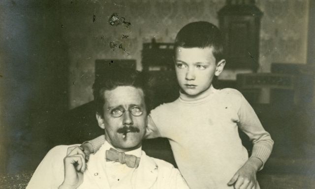 James Joyce is sitting on a chair. His young son has his arm around his father's neck and they are holding hands.